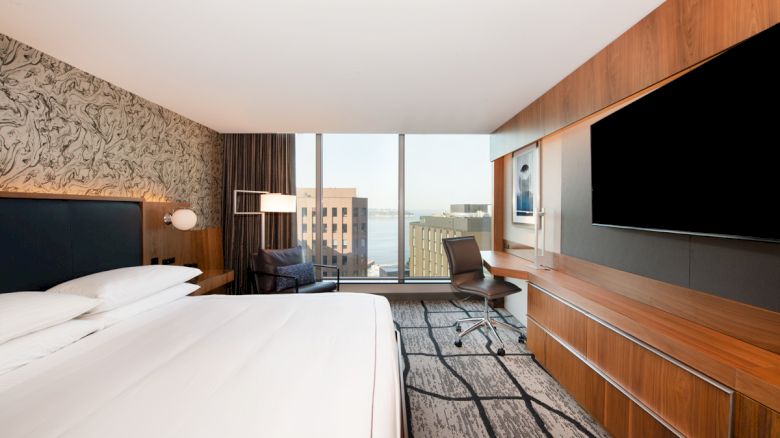 A modern hotel room with a large bed, TV, desk, chair, and a view of buildings and water through a wide window.