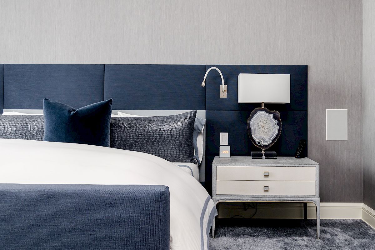 A modern bedroom with a blue upholstered bed, gray carpet, and a nightstand featuring a lamp, decorative object, and wall light above.