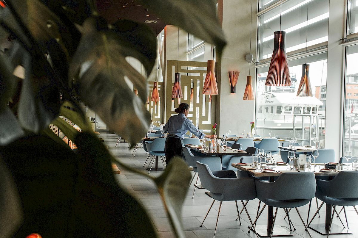 A person sets up tables in a modern restaurant with stylish chairs, pendant lights, and large windows.