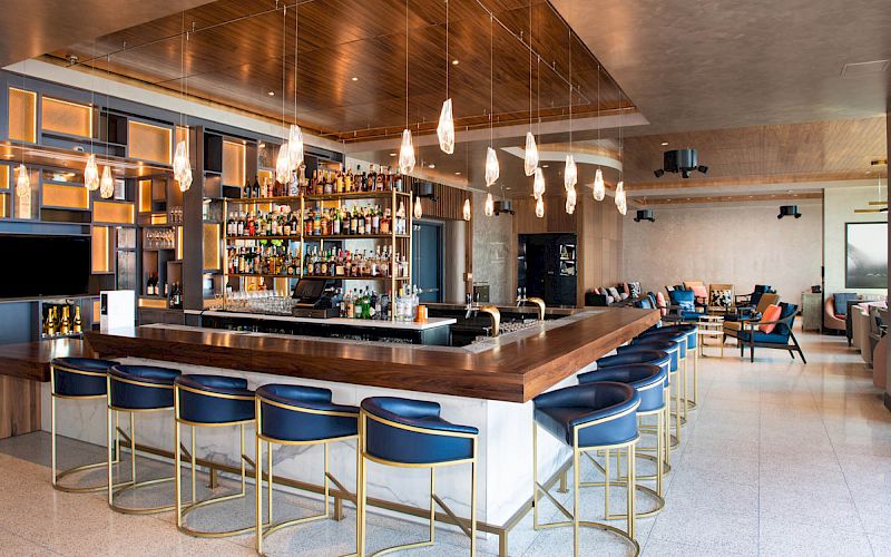 This image shows a modern bar with a wooden counter, blue bar stools, hanging lights, and a well-stocked liquor shelf in a stylish space.