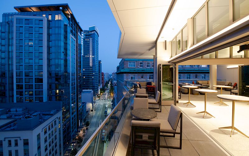 A modern cityscape at dusk with buildings and a furnished balcony featuring chairs and tables, illuminated by soft lighting.