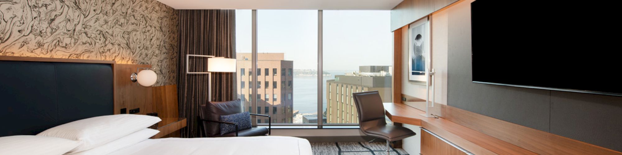 A modern hotel room with a large bed, wall-mounted TV, desk, and window view of city buildings and water.